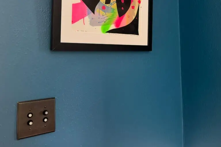 2 gang push button light switch with solid brass bronze-finish switch cover on a blue plaster wall with framed modern art hanging adjacent