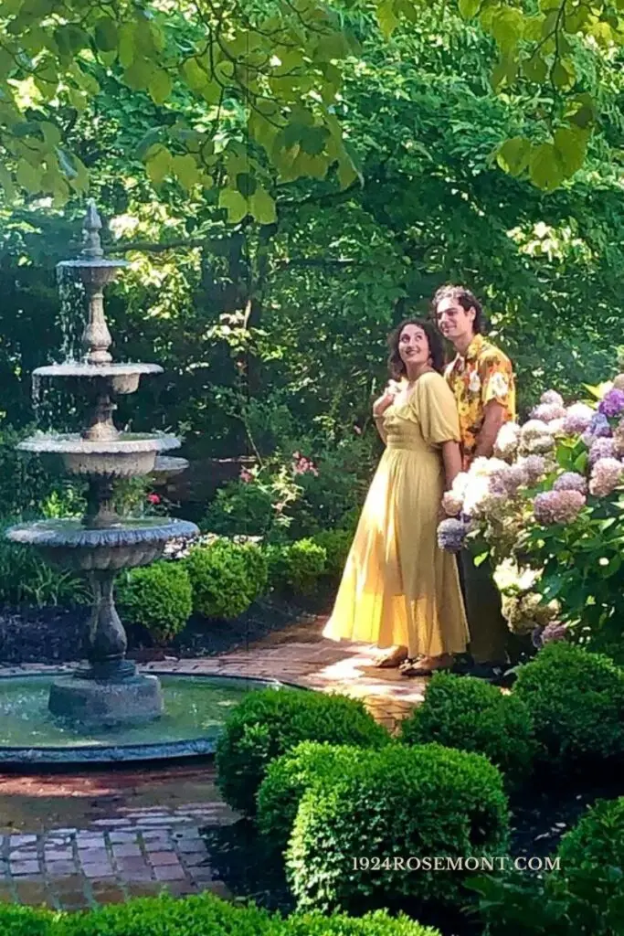 A couple posing for engagement photos beside the three-tier stone fountain on the brick path in the Rosemont garden.