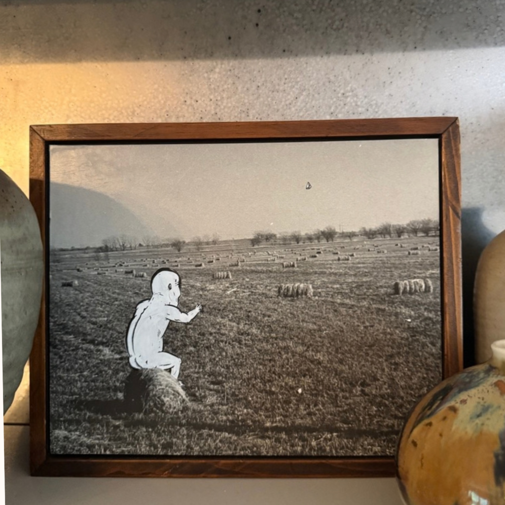 Vintage black-and-white photograph painted with a white figure on a hay bale, leaning on a shelf between studio pottery